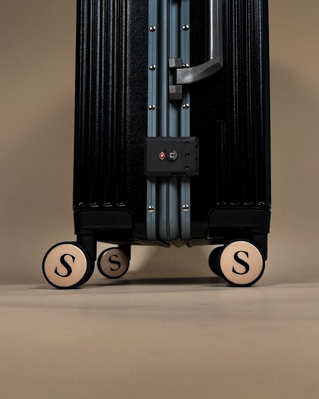 Gros plan sur la base de la valise L’Unique montrant les roues pivotantes beige avec le logo S et un cadenas TSA intégré sur la structure métallique.